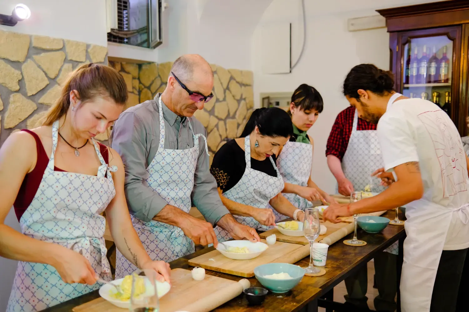 A group of diverse participants wearing aprons learning to make fresh pasta from an instructor during a hands-on Italian cooking class in Rome, with pasta-making ingredients and tools on the wooden counter.