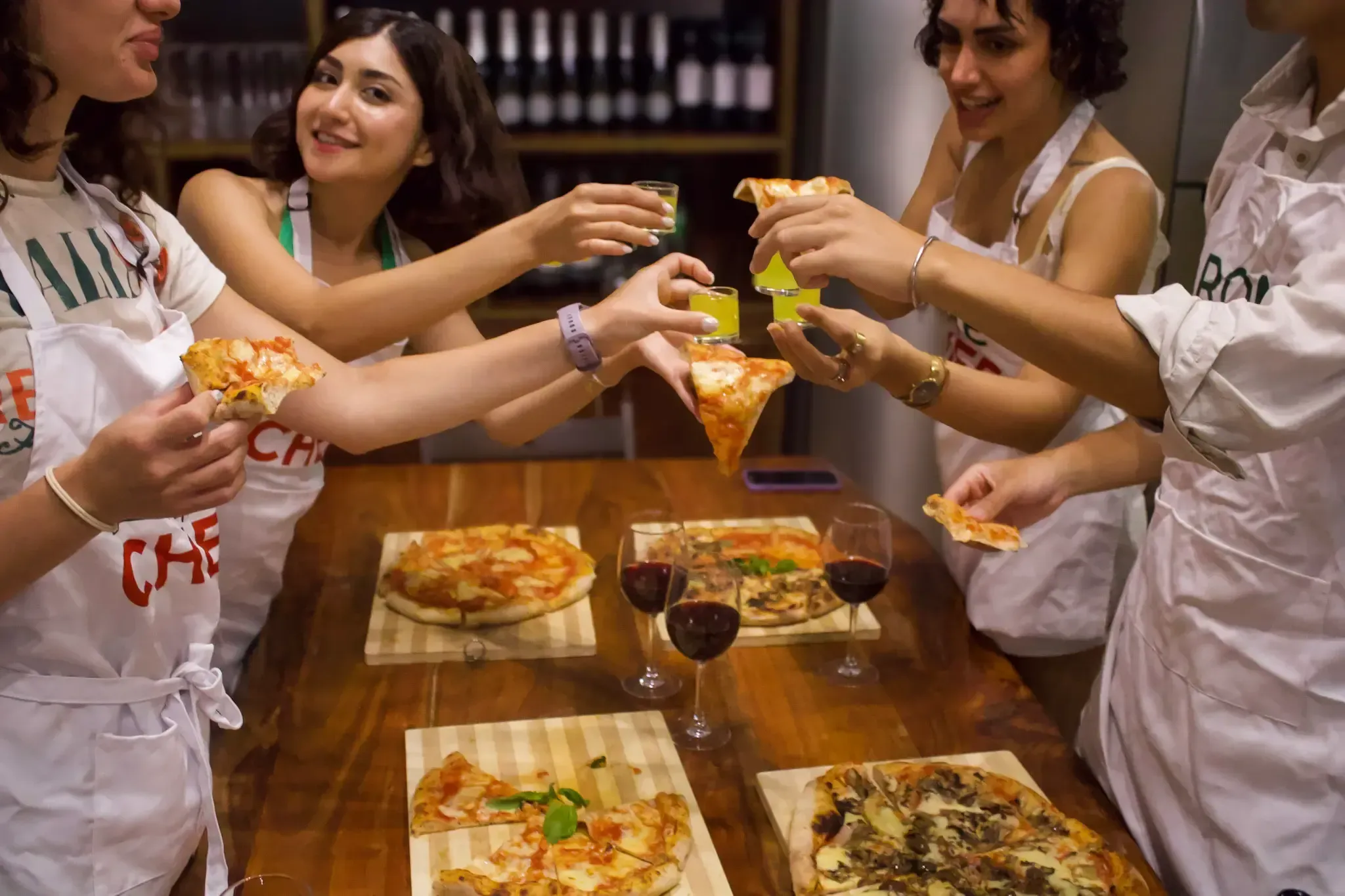 Group of four smiling women in Rome with Chef aprons toasting with wine glasses and holding slices of freshly made pizza